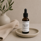 A 1 oz amber glass dropper bottle of The Crystal Garden Clear Skin Face Oil, resting on a small beige ceramic dish. The background features a neutral beige wall, a beige textured cloth, and a blurred vase with green leaves.
