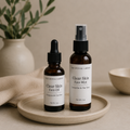 Two amber skincare bottles from The Crystal Garden, a dropper bottle of Face Oil and a spray bottle of Face Mist, displayed on a beige ceramic tray with a neutral background and soft-focus props.