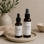 Two amber skincare bottles from The Crystal Garden, a dropper bottle of Face Oil and a spray bottle of Face Mist, displayed on a beige ceramic tray with a neutral background and soft-focus props.