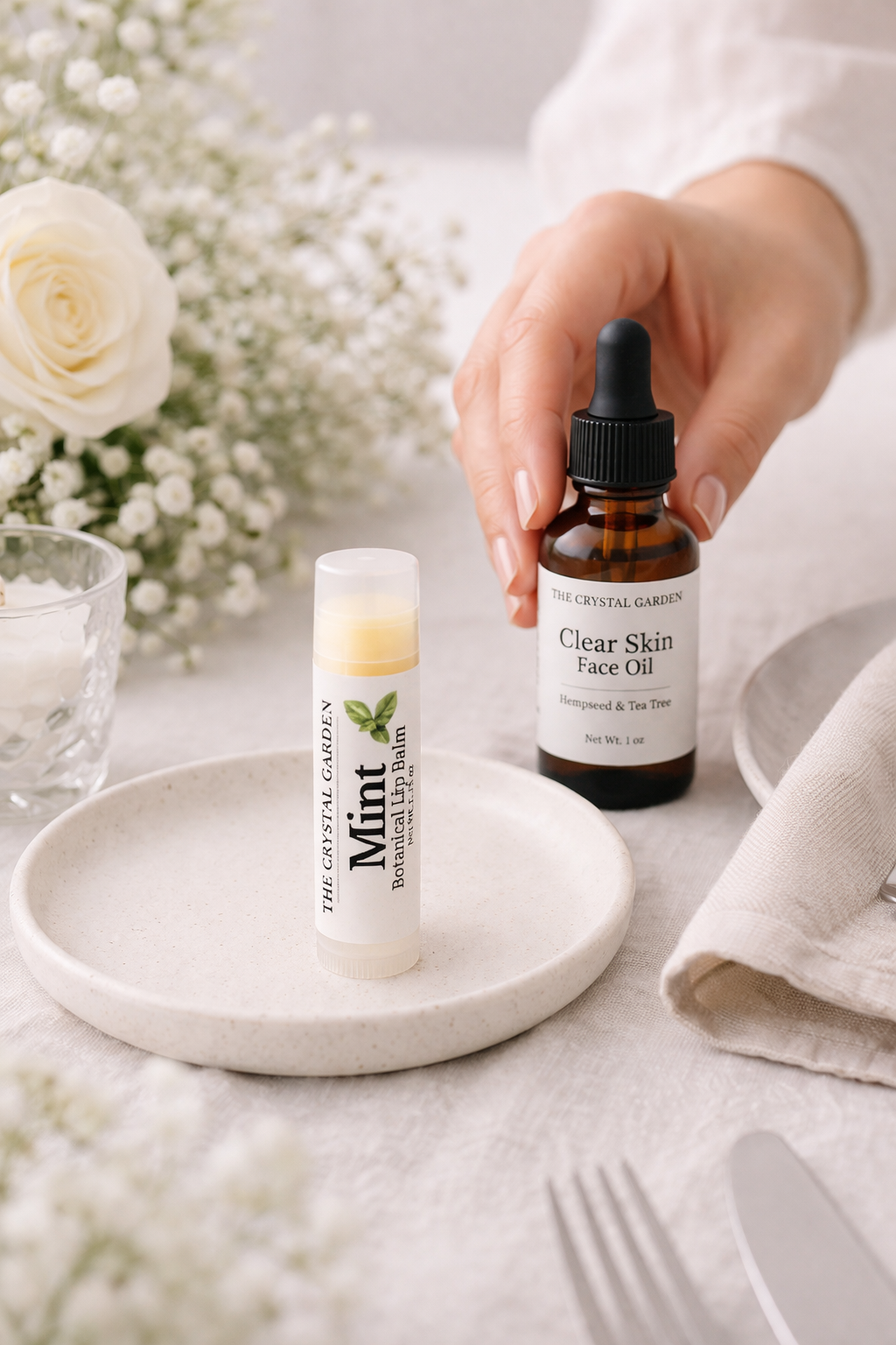 A hand holds a bottle of Clear Skin Face Oil by The Crystal Garden, while a lip balm tube labeled Mint sits on a ceramic dish. The scene is styled with white flowers, a folded linen napkin, and silverware on a light tablecloth.