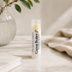 A tube of Cocoa Butter lip balm by The Crystal Garden stands on a light stone surface, with a softly blurred glass vase, green leaves, and a beige knit fabric in the background.