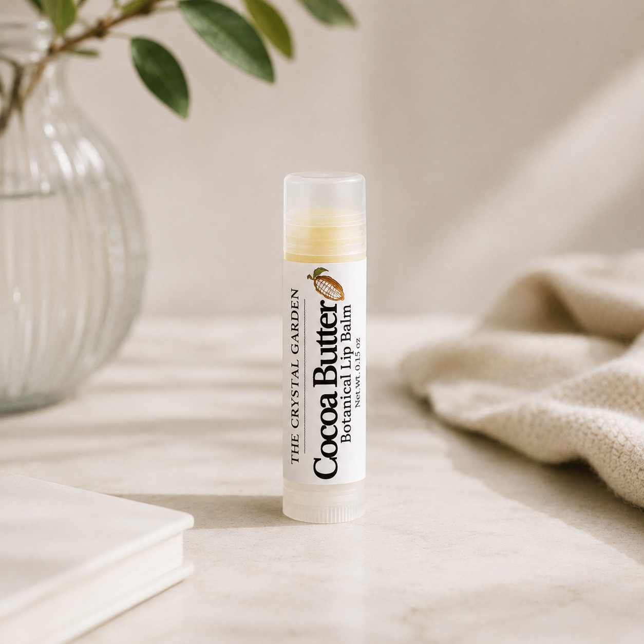 A tube of Cocoa Butter lip balm by The Crystal Garden stands on a light stone surface, with a softly blurred glass vase, green leaves, and a beige knit fabric in the background.