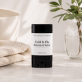 A stick of 'The Crystal Garden' Cold & Flu Botanical Salve stands on a light marble surface. The product has a black container with a white label.