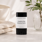 A stick of 'The Crystal Garden' Cold & Flu Botanical Salve stands on a light marble surface. The product has a black container with a white label.