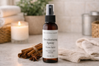 A 2 oz amber glass spray bottle of 'The Crystal Garden' Deodorizing Spray (Home Spice scent) on a marble countertop, styled with cinnamon sticks, a lit candle, and a beige towel.