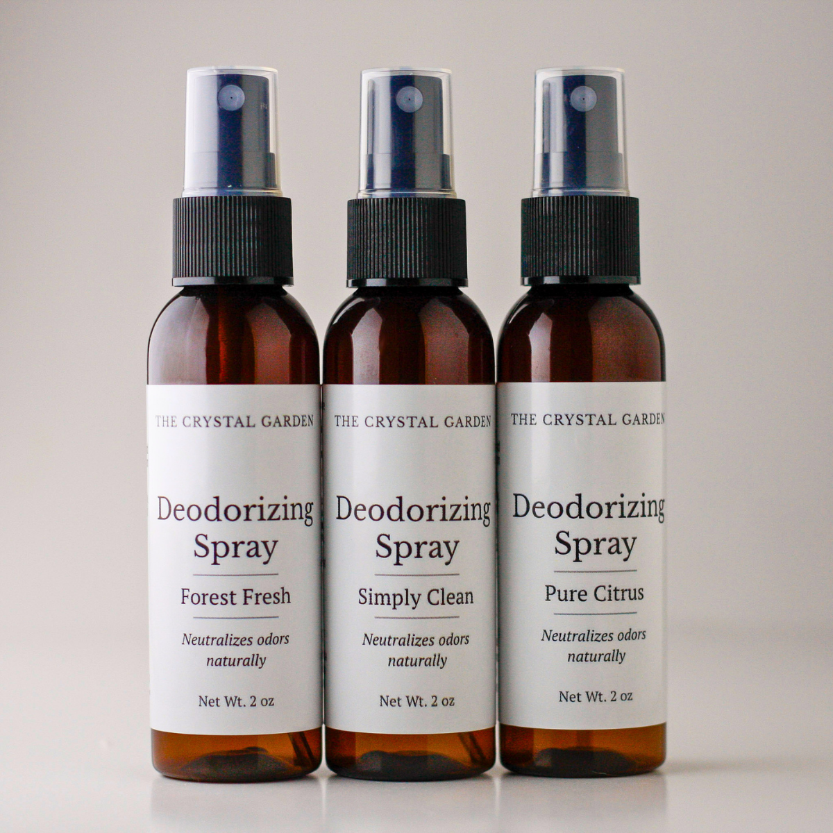 Three amber glass deodorizing spray bottles with white labels and black spray tops, arranged side by side on a light neutral background. The labels read 'The Crystal Garden' and 'Deodorizing Spray' with scent variants Forest Fresh, Simply Clean, and Pure Citrus.