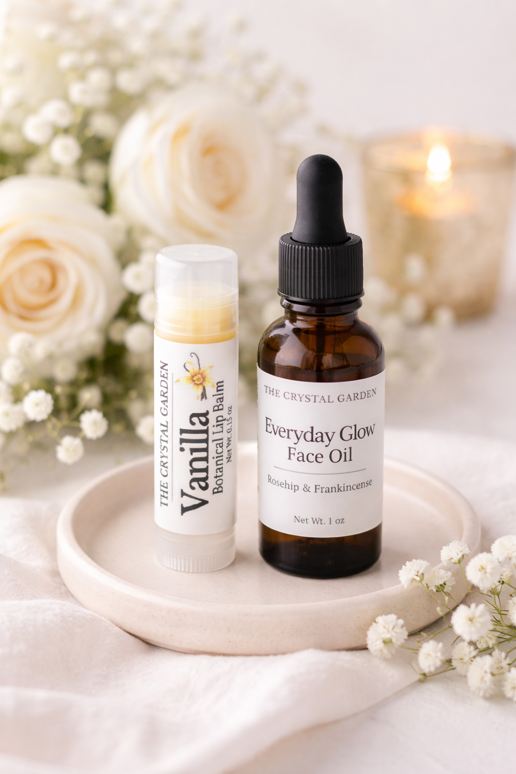 Two skincare products from The Crystal Garden, a Vanilla Botanical Lip Balm and an Everyday Glow Face Oil, displayed on a small white ceramic tray with white roses and baby's breath flowers.