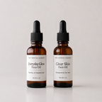 Two amber glass dropper bottles of face oil by The Crystal Garden, labeled Everyday Glow Face Oil and Clear Skin Face Oil, on a light gray background.