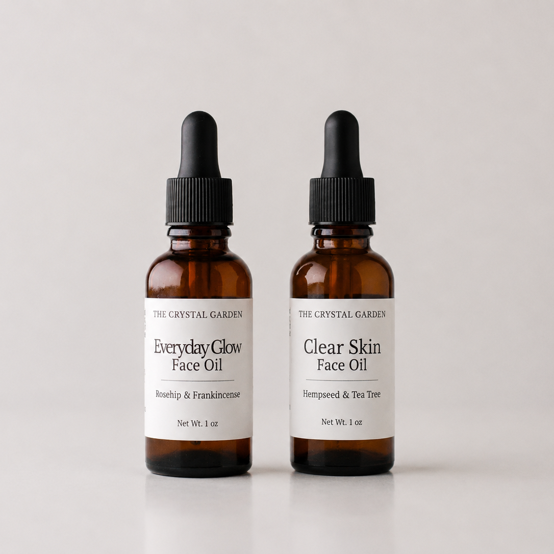 Two amber glass dropper bottles of face oil by The Crystal Garden, labeled Everyday Glow Face Oil and Clear Skin Face Oil, on a light gray background.