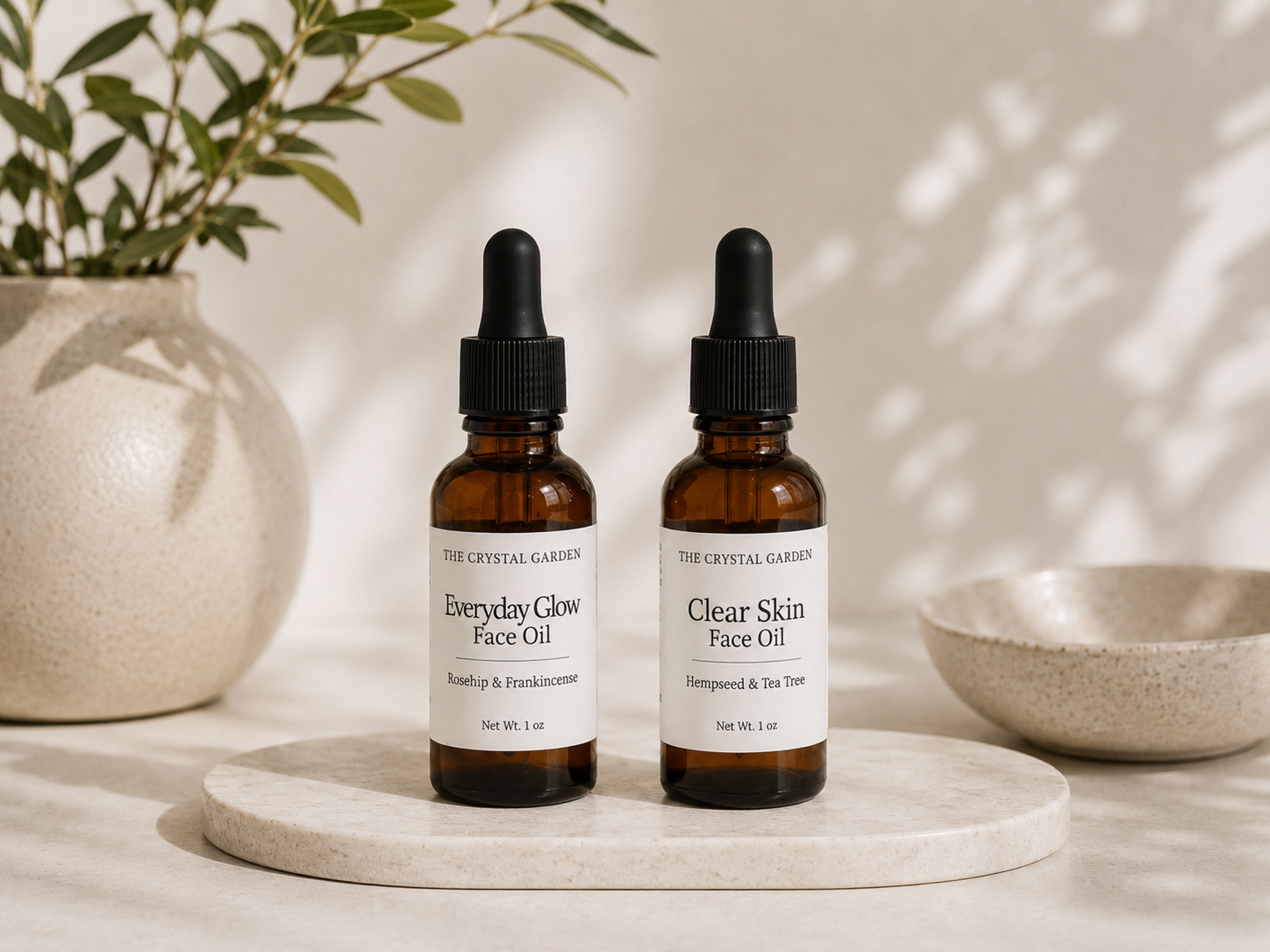 Two amber glass dropper bottles of face oil from The Crystal Garden, labeled Everyday Glow Face Oil and Clear Skin Face Oil, displayed on a round stone tray with a neutral background and soft leaf shadows.
