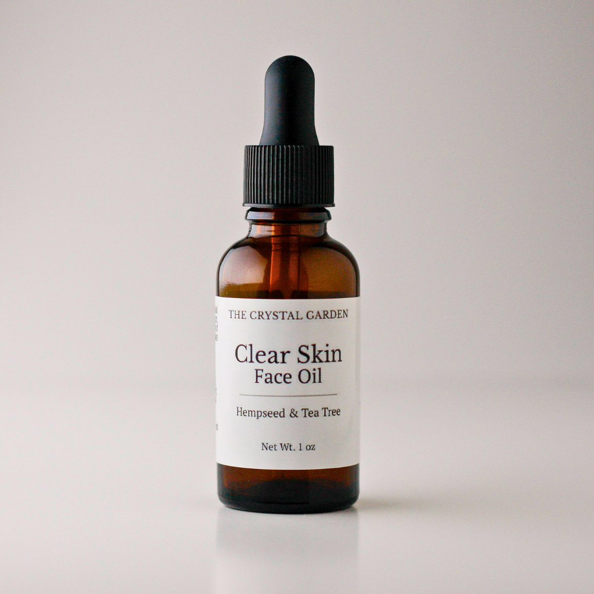 An amber glass dropper bottle of face oil with a black dropper cap and a white label reading 'THE CRYSTAL GARDEN Clear Skin Face Oil Hempseed & Tea Tree'.