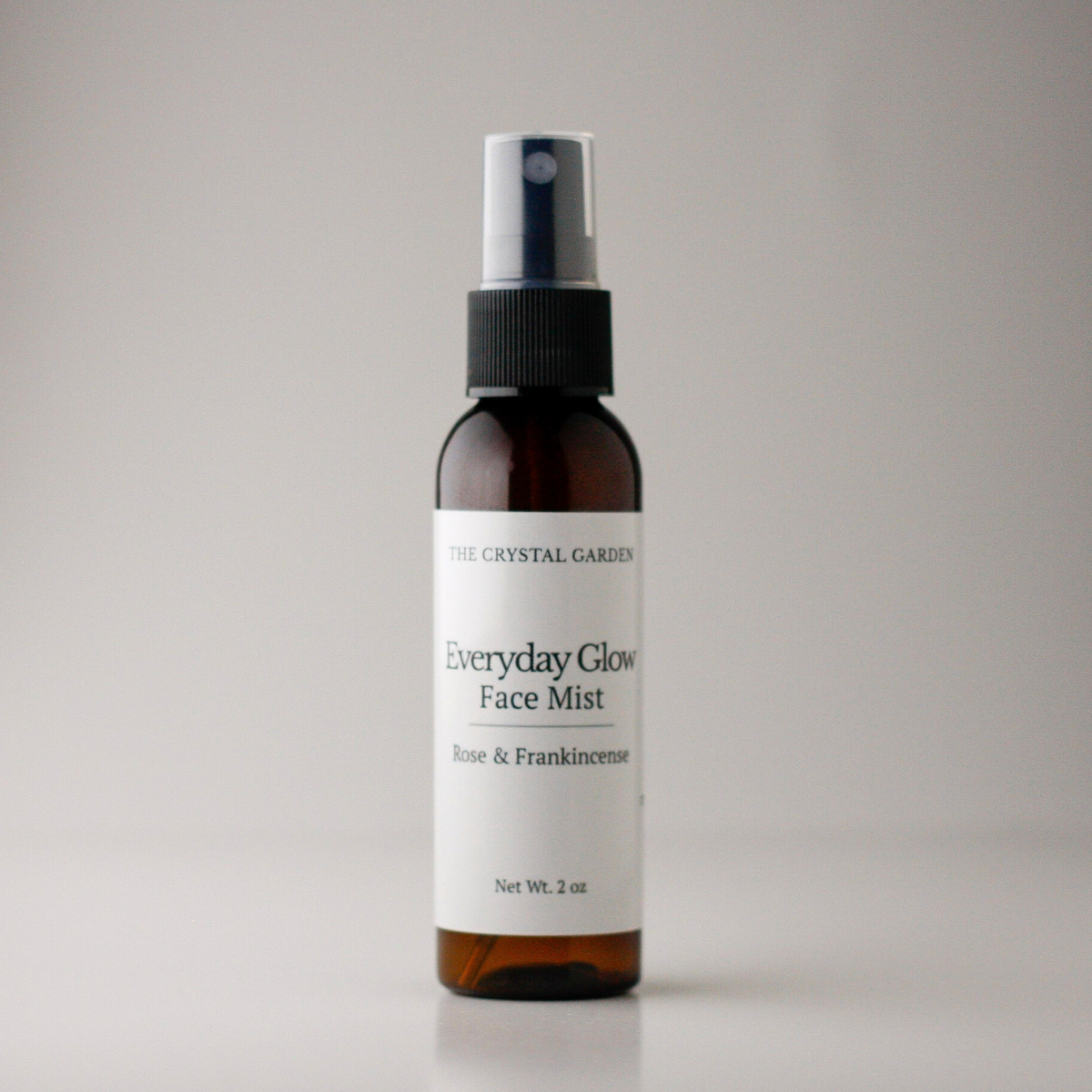A 2 oz amber spray bottle of The Crystal Garden Everyday Glow Face Mist with Rose & Frankincense, featuring a white label and black spray nozzle, set against a neutral gray background.