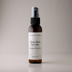 A 2 oz amber glass spray bottle of The Crystal Garden Clear Skin Face Mist with lavender and tea tree, featuring a white label and black spray nozzle, set against a neutral gray background.