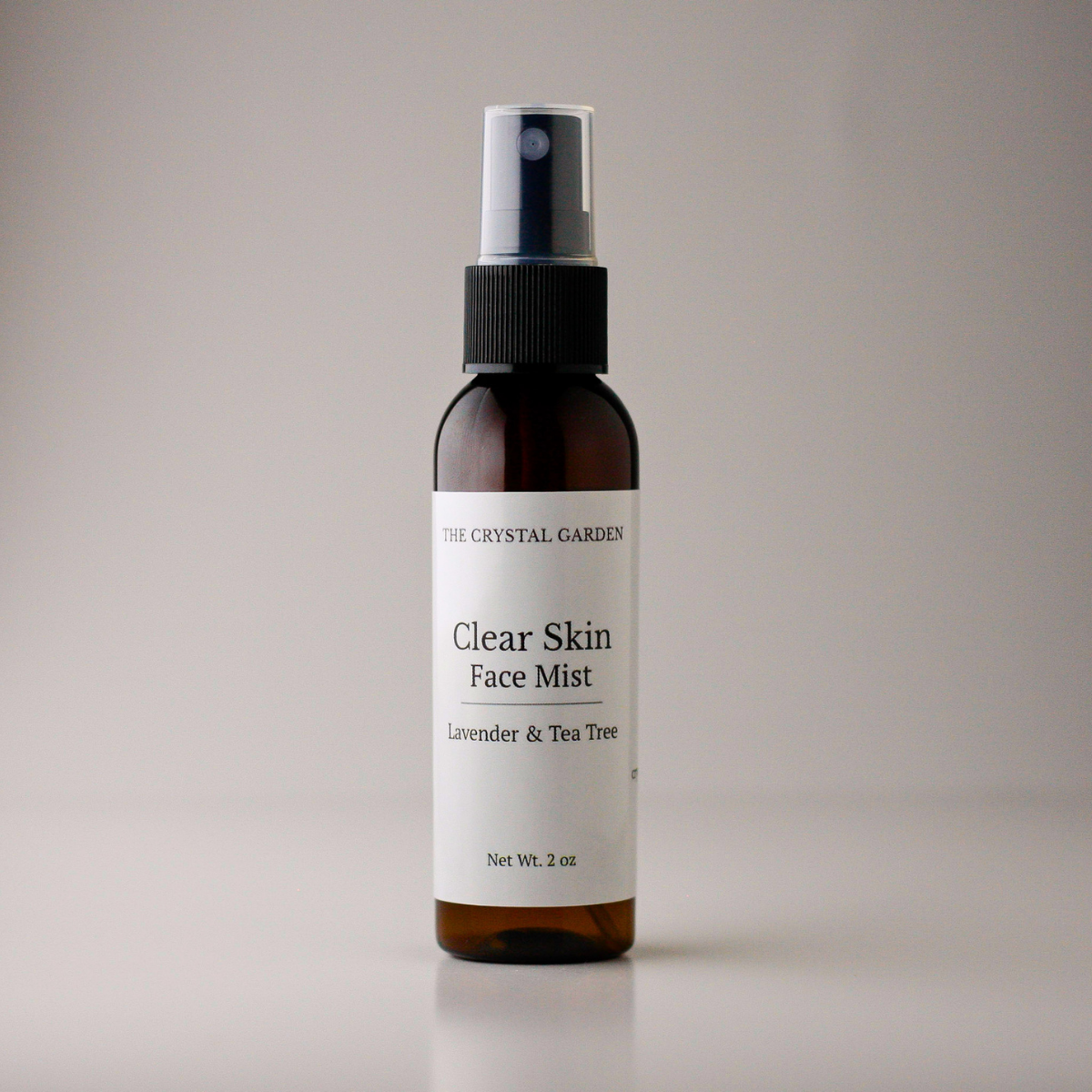 A 2 oz amber glass spray bottle of The Crystal Garden Clear Skin Face Mist with lavender and tea tree, featuring a white label and black spray nozzle, set against a neutral gray background.