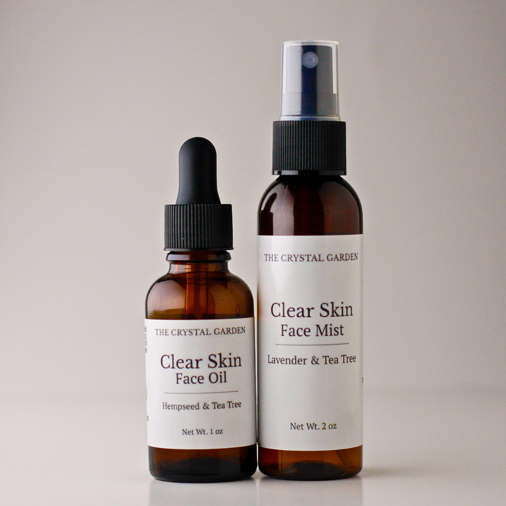Two amber glass bottles from The Crystal Garden on a light gray background: a smaller dropper bottle labeled Clear Skin Face Oil and a larger spray bottle labeled Clear Skin Face Mist with lavender and tea tree.