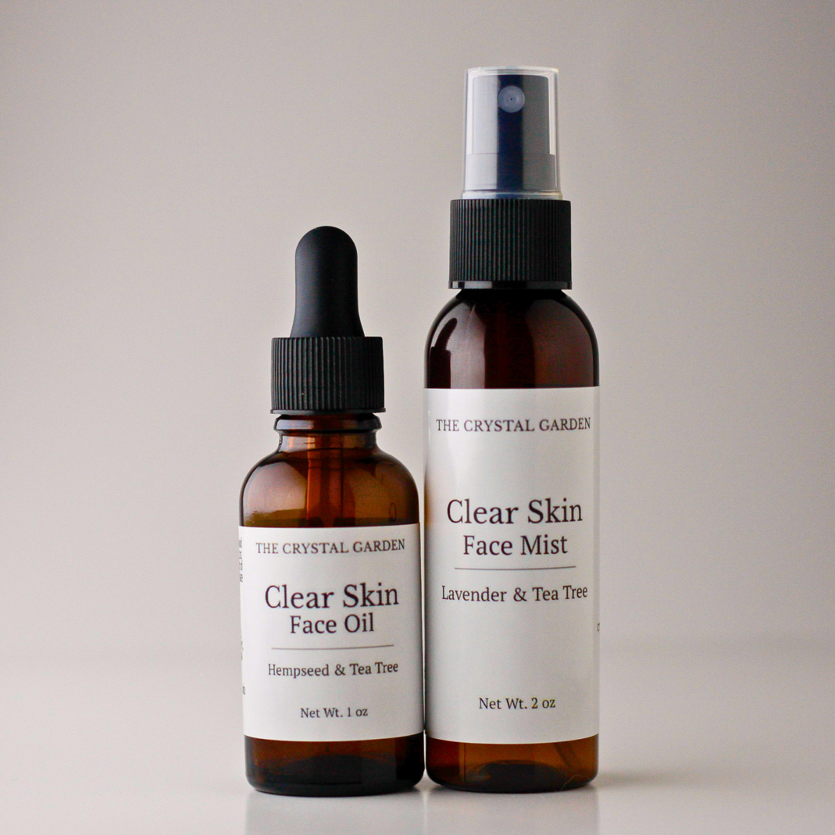 Two amber glass bottles from The Crystal Garden on a light gray background: a smaller dropper bottle labeled Clear Skin Face Oil and a larger spray bottle labeled Clear Skin Face Mist with lavender and tea tree.