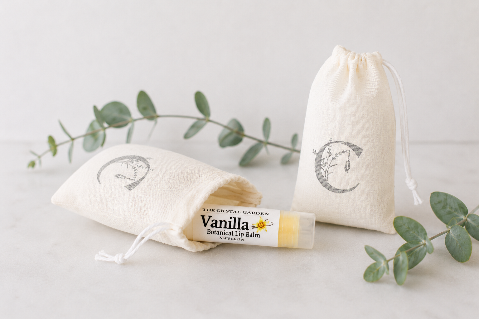 A lip balm tube labeled 'Vanilla Botanical Lip Balm' by The Crystal Carvings, displayed with two cream drawstring pouches on a light surface with eucalyptus sprigs.