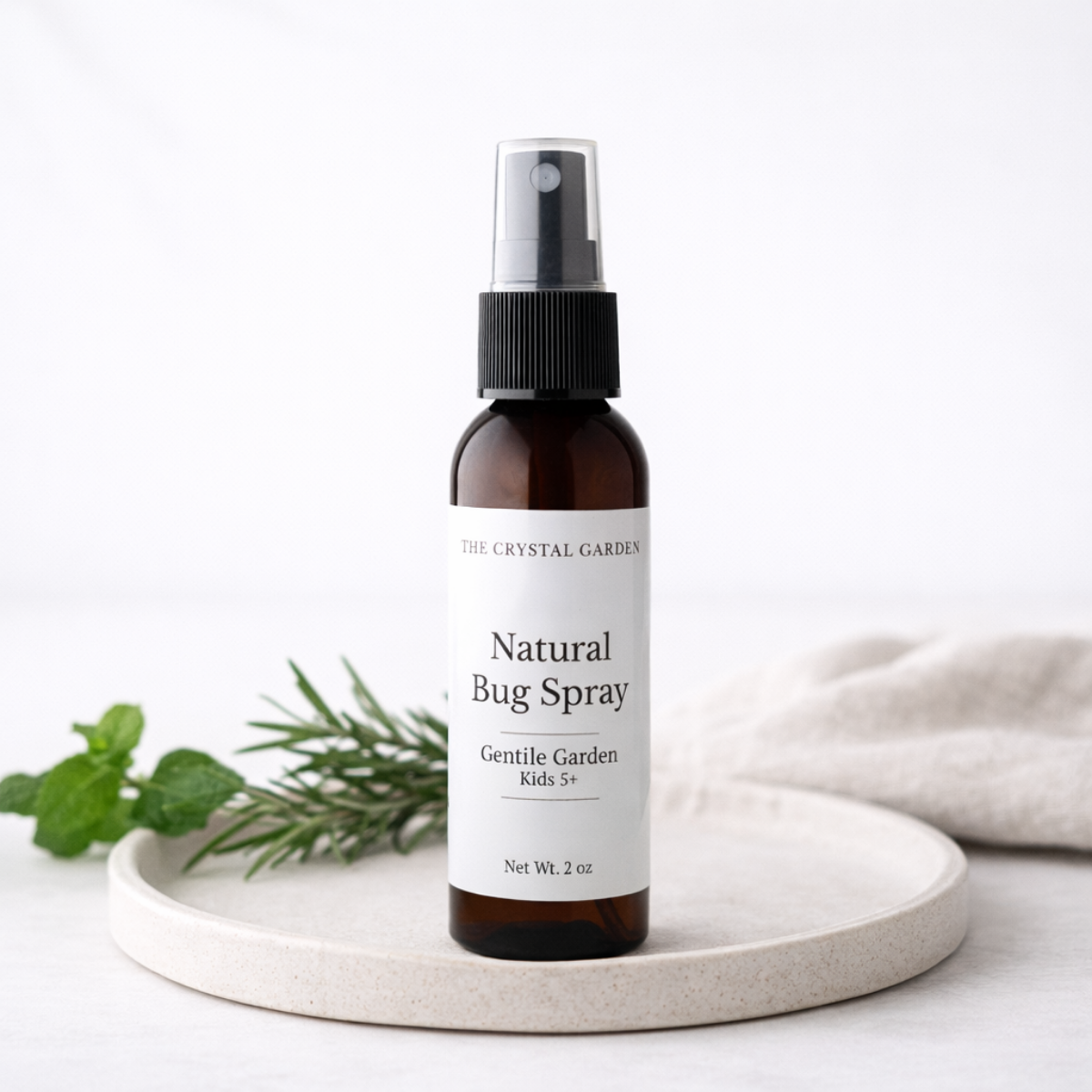 A 2 oz amber glass spray bottle of The Crystal Garden Natural Bug Spray with a white label, styled on a light beige ceramic tray with mint leaves and a soft beige cloth against a white background.