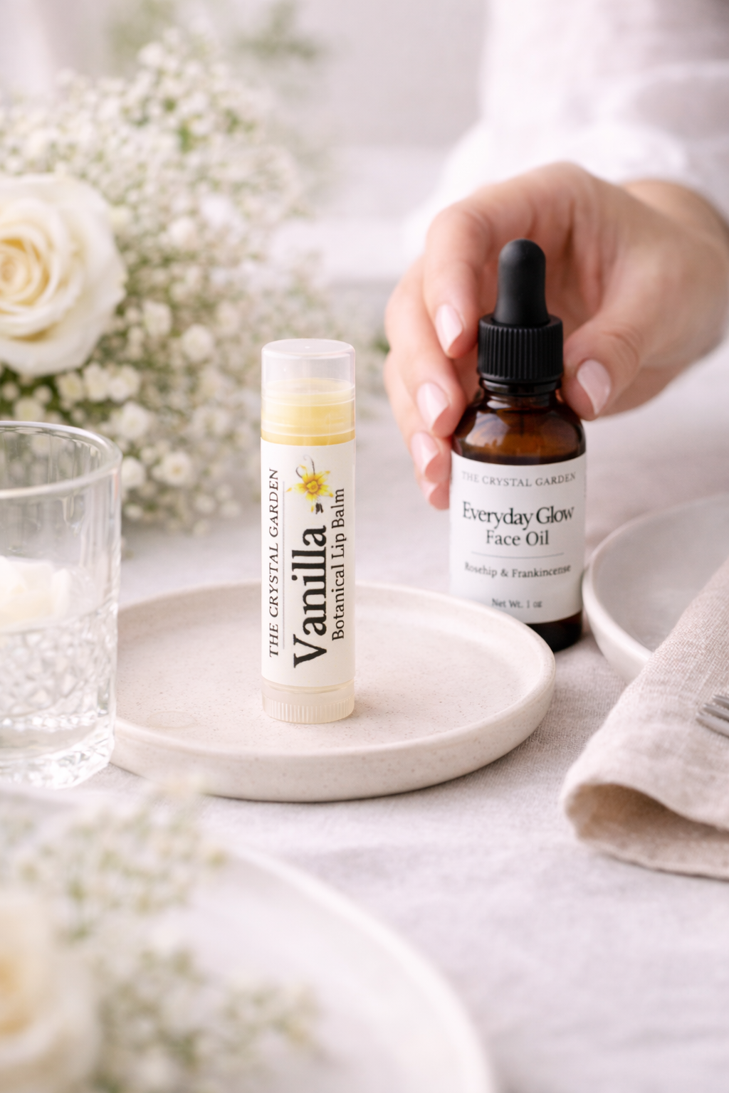 A hand holds an amber dropper bottle of Everyday Glow Face Oil next to a tube of Vanilla Botanical Lip Balm from The Crystal Garden, styled on a light table with flowers and plates.