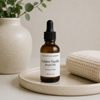 A 1 oz amber glass dropper bottle of 'Golden Vanilla Beard Oil' by The Crystal Garden, styled on a speckled beige ceramic tray next to a rolled cream knit towel and a ceramic vase with green leaves.