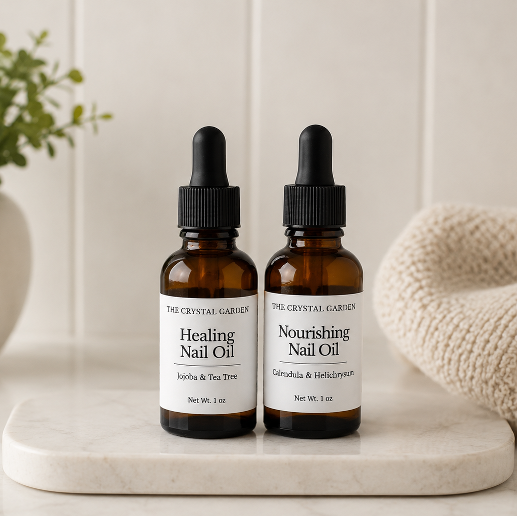 Two amber glass dropper bottles of nail oil labeled 'Healing Nail Oil' and 'Nourishing Nail Oil' by The Crystal Gardens, displayed on a white marble tray with a potted plant and a knitted blanket in the background.