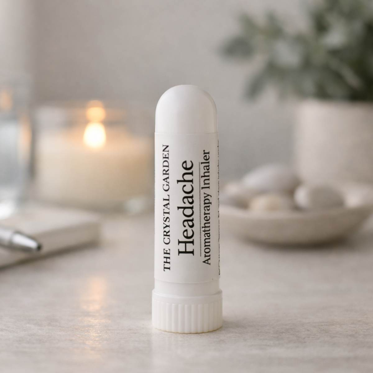 A white cylindrical atomizer tube labeled 'Headache Aromatherapy Inhaler' by The Crystal Garden, standing upright on a light stone surface with a softly blurred candle, pen, and plant in the background.