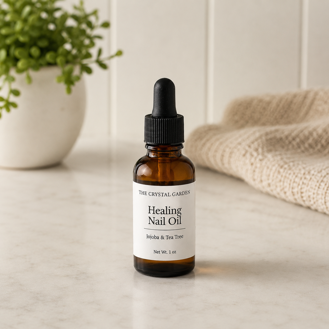 A small amber glass dropper bottle labeled 'THE CRYSTAL GARDEN Healing Nail Oil' sits on a light marble surface with a blurred potted plant and beige textile in the background.