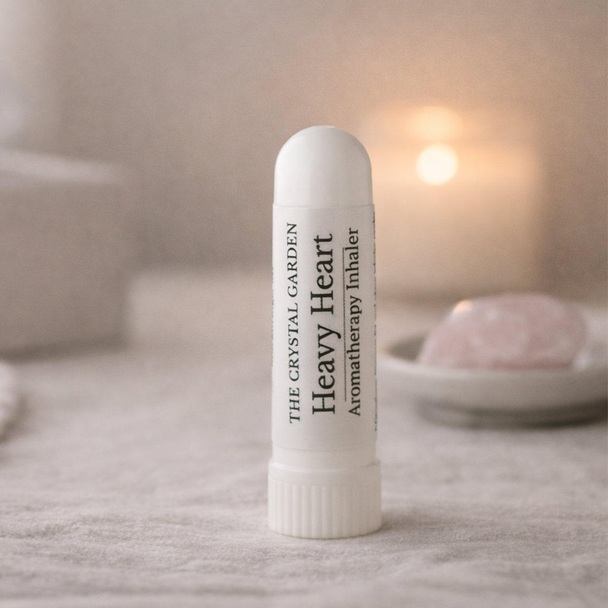 A white lip balm tube labeled 'THE CRYSTAL GARDEN Heavy Heart Aromatherapy Inhaler' standing upright on a soft fabric surface with a blurred warm background and a pink stone in the distance.