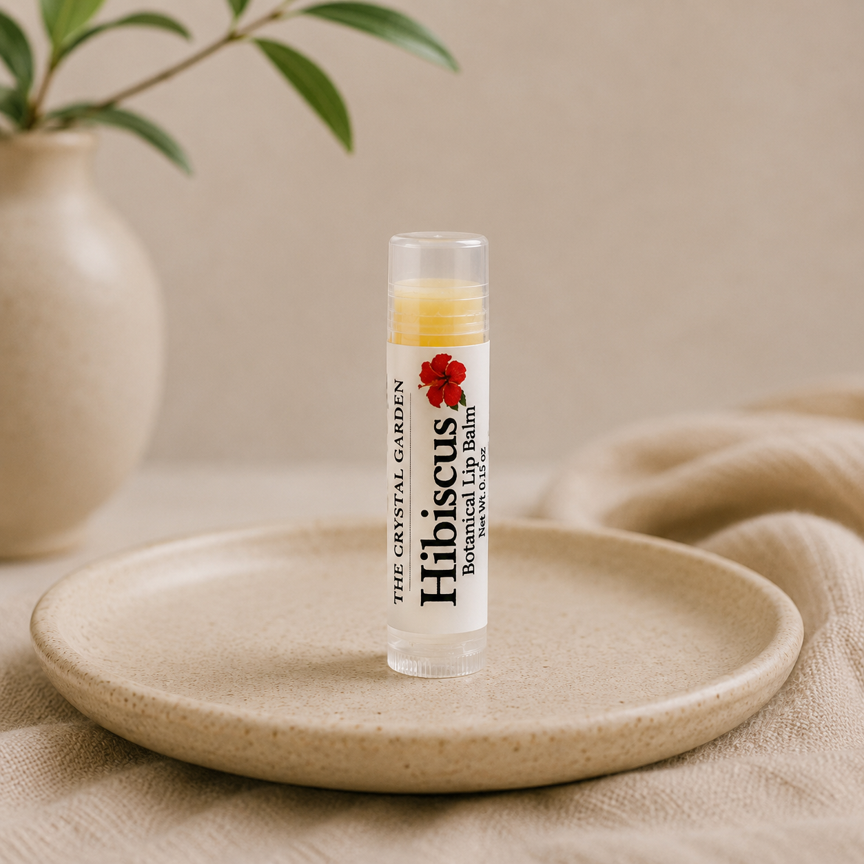 A clear lip balm tube standing upright on a round beige ceramic plate, with a soft neutral fabric backdrop and a blurred vase of green leaves in the background. The label reads 'THE CRYSTAL GARDEN Hibiscus Botanical Lip Balm' with a red hibiscus flower graphic.