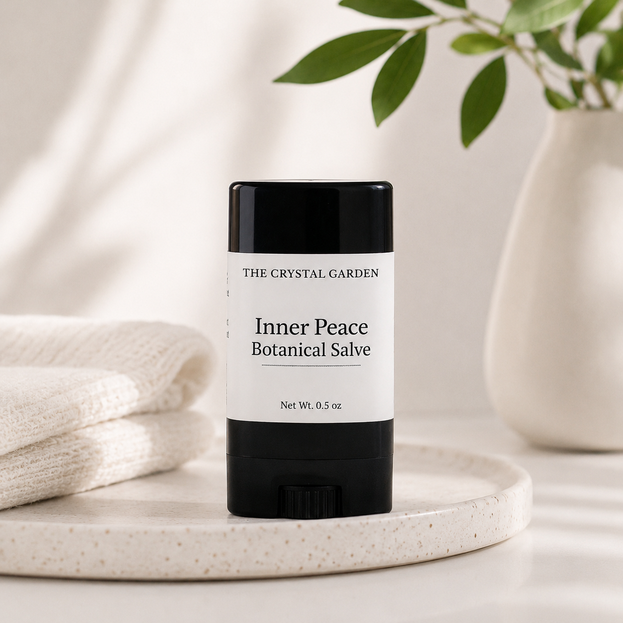 A black and white stick of 'Inner Peace Botanical Salve' by The Crystal Garden, styled on a speckled ceramic tray with a folded cream towel and a white vase in the background.