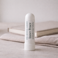 A white cylindrical aromatherapy inhaler tube labeled 'The Crystal Garden Inner Peace Aromatherapy Inhaler' standing upright on a light surface with folded fabric and a book blurred in the background.