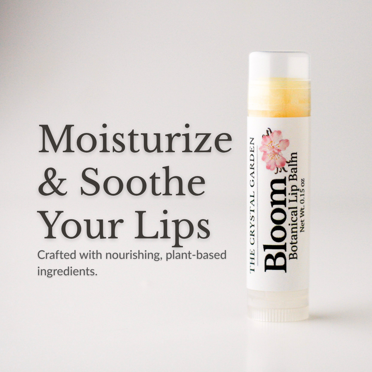 A tube of Bloom Botanical Lip Balb by The Crystal Garden stands on a light gray background next to large text that reads 'Moisturize & Soothe Your Lips'. The lip balm tube is white with a clear cap, showing a pale yellow balm inside, and has a pink floral graphic on the label.