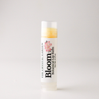 A single tube of lip balm from The Crystal Garden's Bloom Botanical Lip Balm, featuring a white label with a pink floral graphic and black text, set against a plain light gray background.