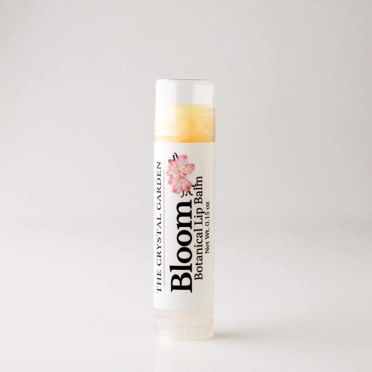 A single tube of lip balm from The Crystal Garden's Bloom Botanical Lip Balm, featuring a white label with a pink floral graphic and black text, set against a plain light gray background.