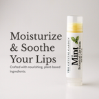 A tube of mint botanical lip balm from The Crystal Garden stands on a white surface against a light gray background, next to promotional text that reads 'Moisturize & Soothe Your Lips.'.