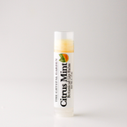 A tube of 'Citrus Mint Botanical Lip Balm' by 'The Crystal Garden' standing upright on a light gray background.