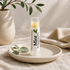 A tube of mint botanical lip balm by The Crystal Garden sits on a speckled ceramic dish, styled with eucalyptus leaves and a neutral linen cloth.