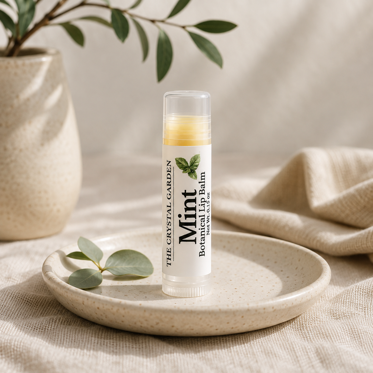 A tube of mint botanical lip balm by The Crystal Garden sits on a speckled ceramic dish, styled with eucalyptus leaves and a neutral linen cloth.