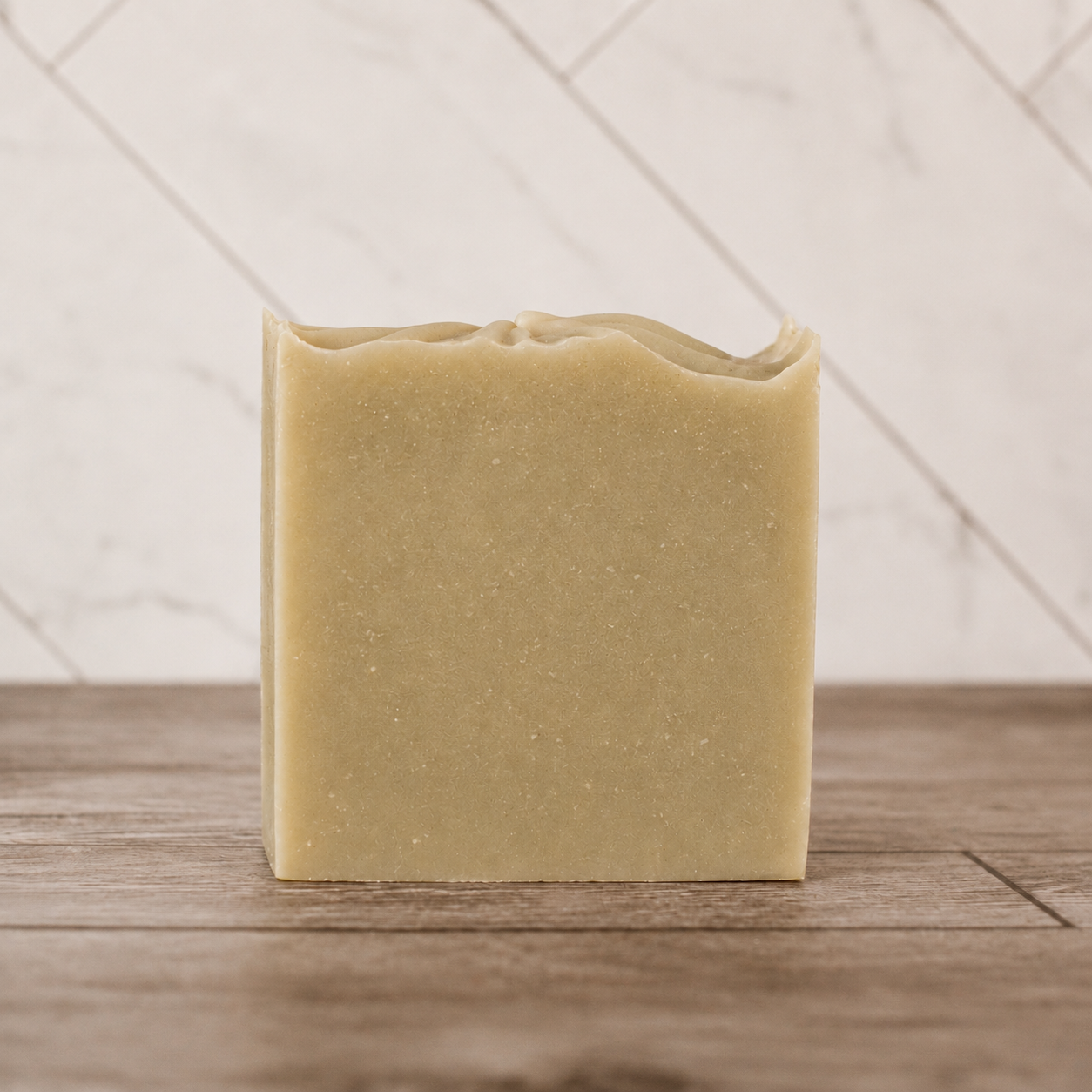 A single rectangular bar of pale yellow-green soap with a rustic, uneven top edge, standing upright on a wooden surface against a light tiled wall.