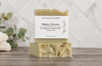 A bar of botanical soap labeled 'Misty Grove' by The Crystal Garden, stacked on another soap bar on a wooden surface with a blurred background of eucalyptus leaves and white flowers.