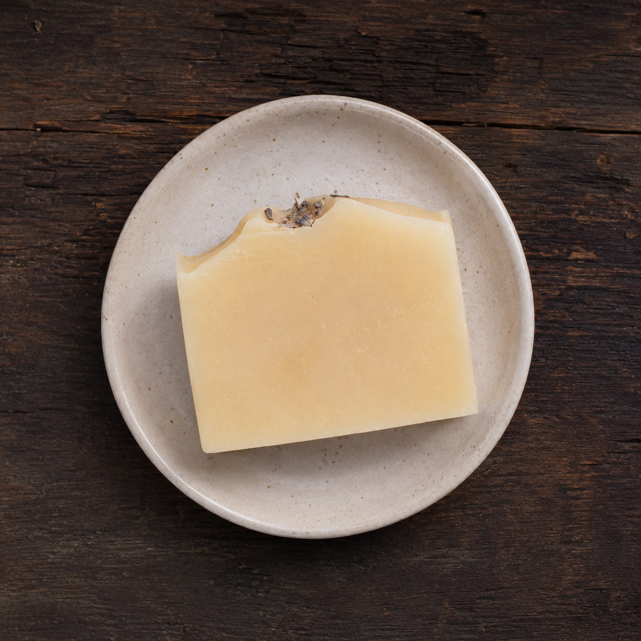 A rectangular bar of pale yellow soap with a small dried herb sprig on top, resting on a round speckled beige ceramic plate over a dark rustic wooden surface.