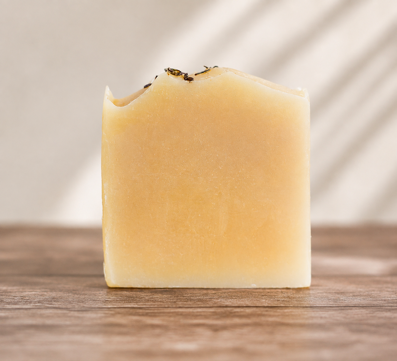 A single rectangular bar of pale yellow soap with a rustic, uneven top edge and small dried herb pieces on top, standing on a wooden surface against a softly blurred background with diagonal light shadows.