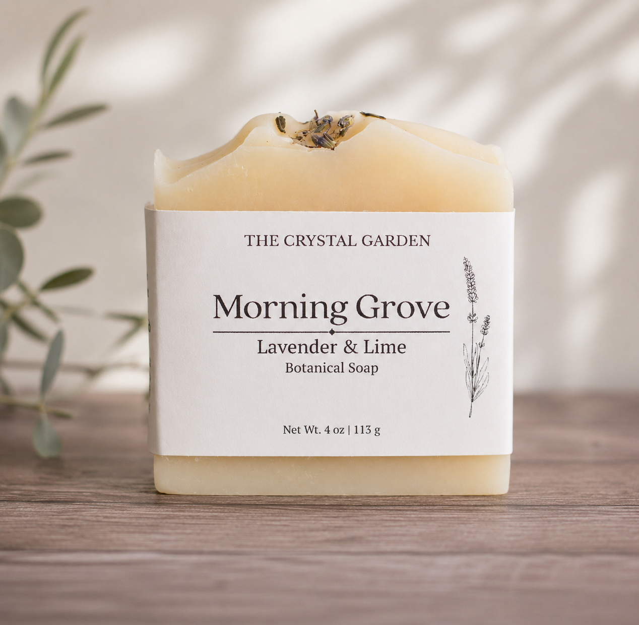 A bar of 'Morning Grove' botanical soap by The Crystal Garden, featuring a white label and a pale yellow wax body with dried lavender buds on top, displayed on a wooden surface.