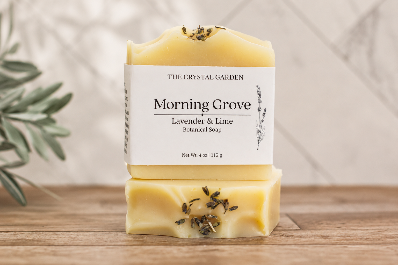 Two stacked bars of The Crystal Garden Morning Grove Lavender & Lime Botanical Soap on a wooden surface, with a soft-focus background and green leaves on the left.