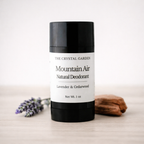 A 1 oz stick deodorant from The Crystal Garden in the Mountain Air scent, featuring a black container with a white label, styled on a light wood surface with lavender and a piece of wood in the background.