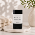 A black and white stick of 'The Crystal Garden' Muscle Ease Botanical Salve sits on a speckled ceramic dish, styled with a folded beige knit blanket on the left and a ceramic vase with green leaves on the right.