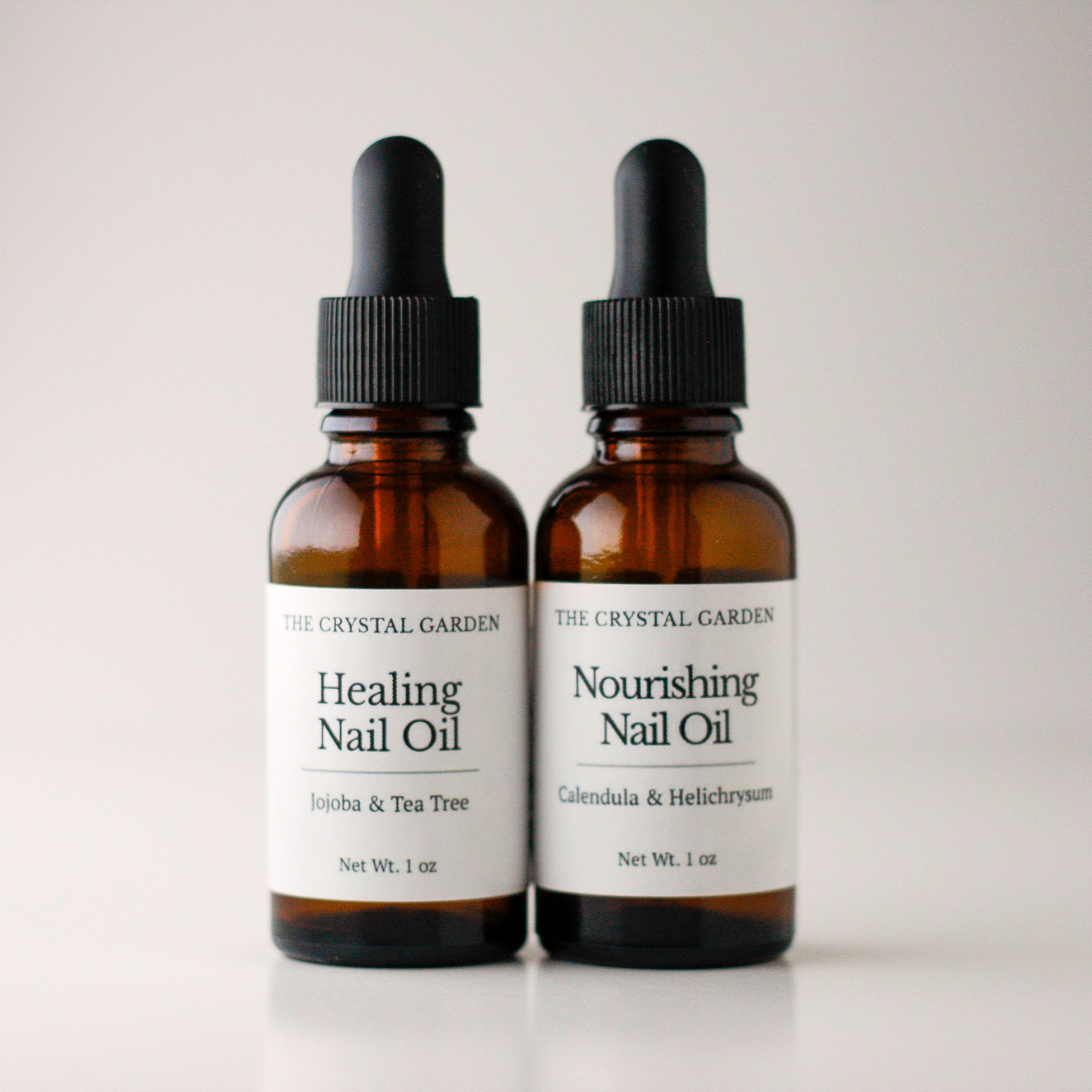 Two amber glass dropper bottles of nail oil from The Crystal Garden, one labeled Healing Nail Oil with Jojoba & Tea Tree and the other Nourishing Nail Oil with Calendula & Helichrysum, set against a clean light gray background.
