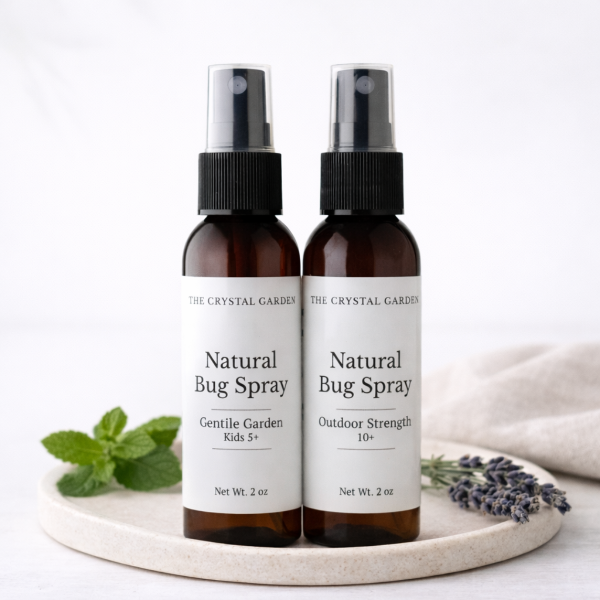 Two amber spray bottles of The Crystal Garden Natural Bug Spray with white labels, displayed on a round stone tray with mint leaves and lavender sprigs against a clean white background.