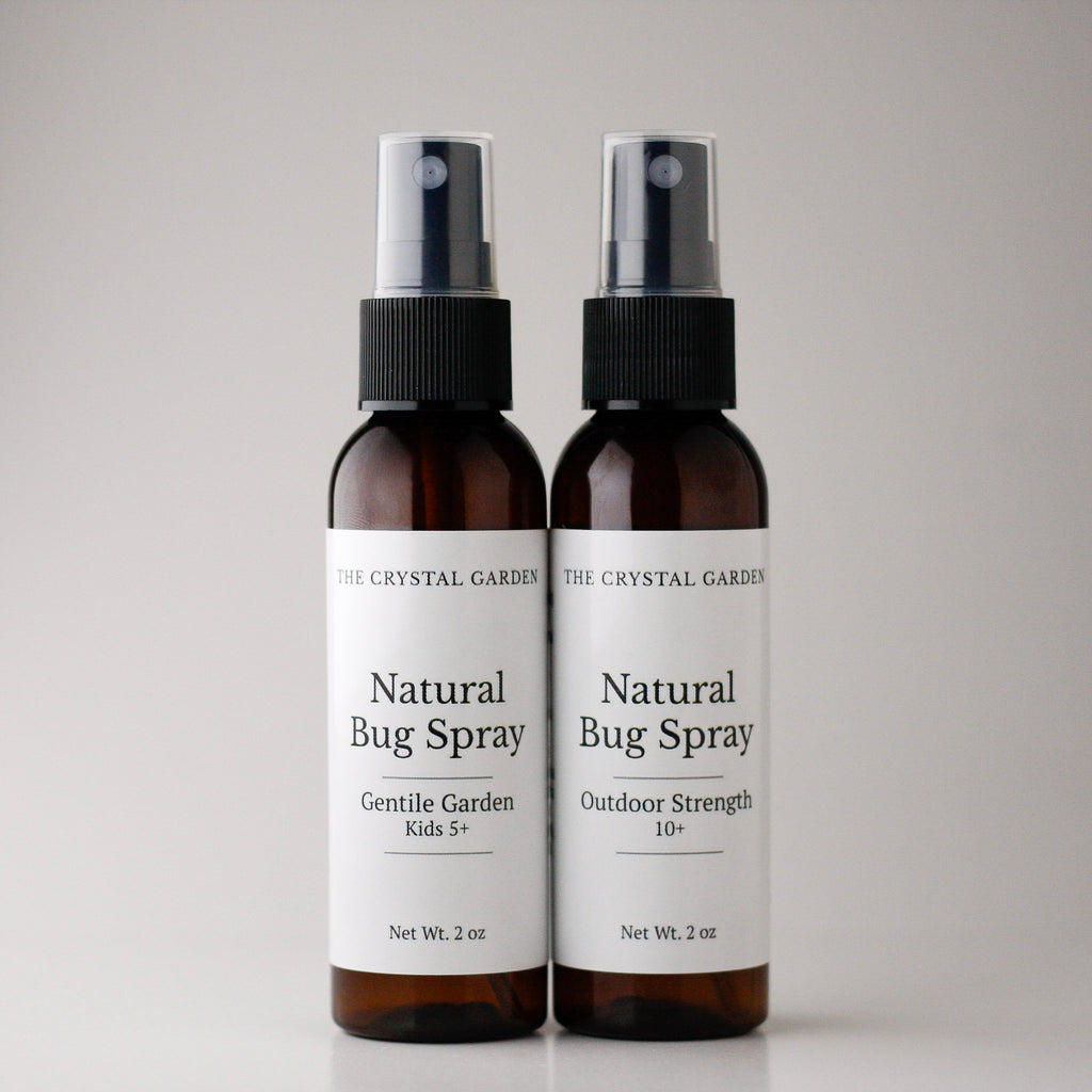 Two amber spray bottles of The Crystal Garden Natural Bug Spray standing side by side on a light gray background.