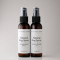 Two amber spray bottles of The Crystal Garden Natural Bug Spray standing side by side on a light gray background.